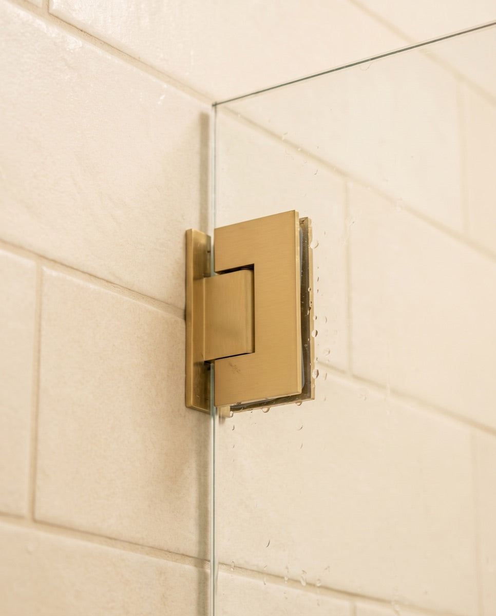 A detail of a frameless glass shower door corner with a slim brushed-gold hinge.