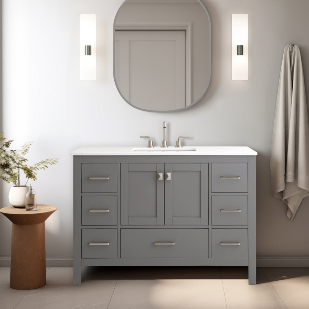 The Vanea 48 inch Bathroom Vanity in Gray with White Quartz Top sits below an oval mirror, flanked by two wall lights. A wooden stool with a vase of flowers and a towel adds charm beside the vanity.