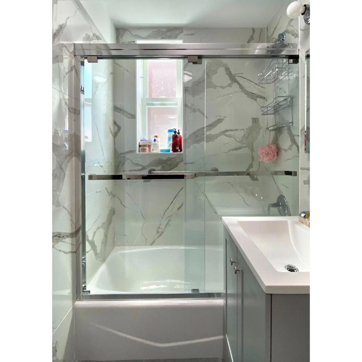 Modern bathroom featuring a white bathtub behind the ASD Cascade Shower Door – Bypass Sliding, marble-patterned walls, a small window above the tub, and a white sink with gray vanity. Toiletries rest on the tub ledge and shower caddy.