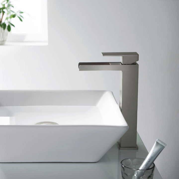 A modern bathroom sink features a rectangular white basin and the Quadra Waterfall Single Handle Bathroom Vessel Sink Faucet. In the background, a small plant sits on the windowsill, with a glass holding toothpaste on the counter.