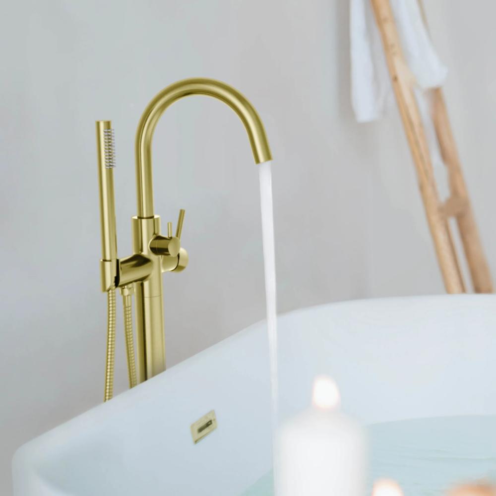The Halo Floor Mount Tub Filler With Hand Shower fills a white tub, while a lit candle and towels on a wooden ladder in the background create a calm, spa-like atmosphere.