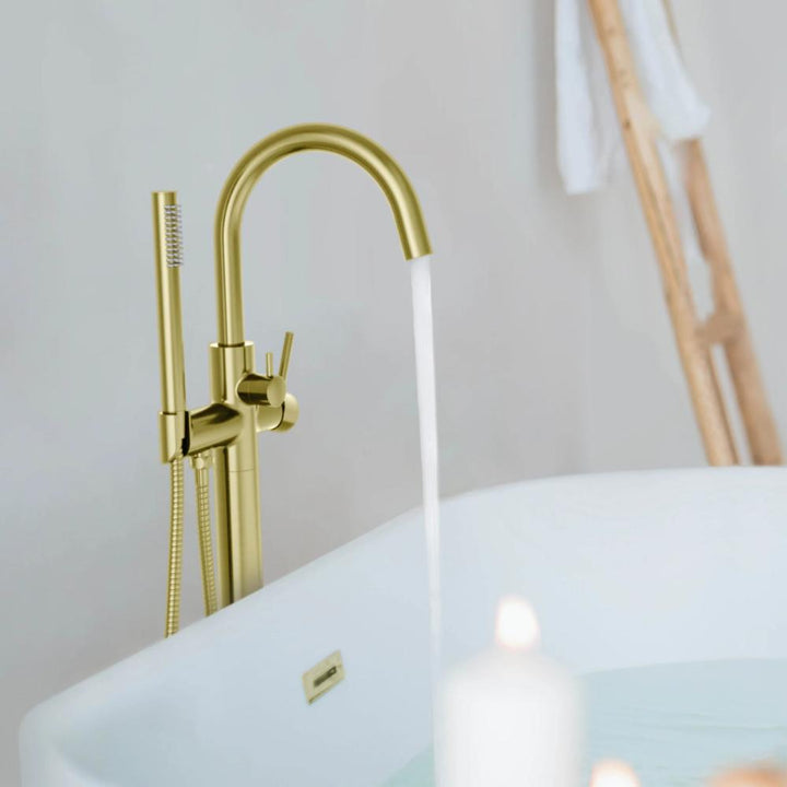 The Halo Floor Mount Tub Filler With Hand Shower fills a white tub, while a lit candle and towels on a wooden ladder in the background create a calm, spa-like atmosphere.
