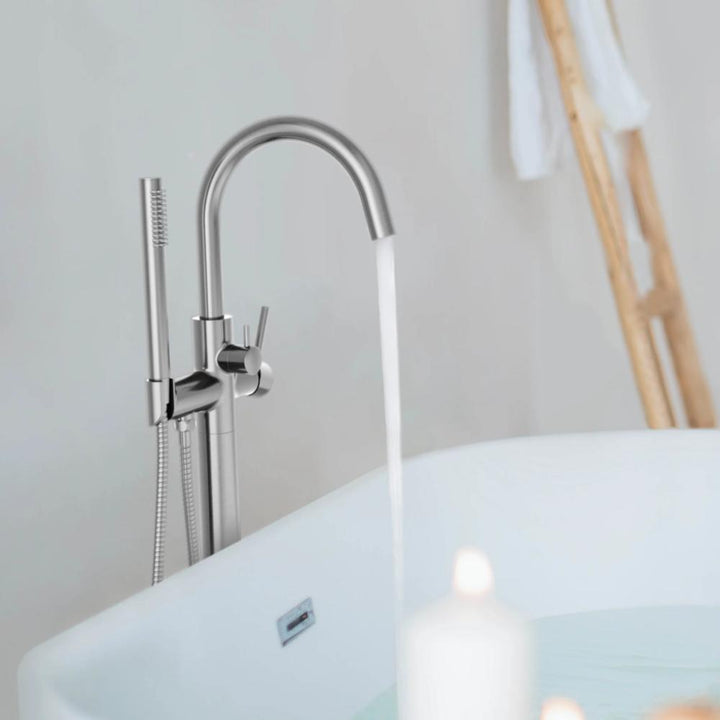 The Halo Floor Mount Tub Filler With Hand Shower stands by a white tub, while a wooden ladder with a towel and a glowing candle help create a tranquil, spa-like setting.