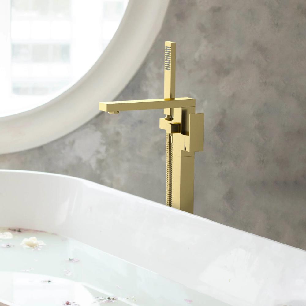 The Quadra Floor Mount Tub Filler With Hand Shower, featuring a modern square gold design, is installed beside a white bathtub filled with water and flower petals, set against a textured gray wall and round window.