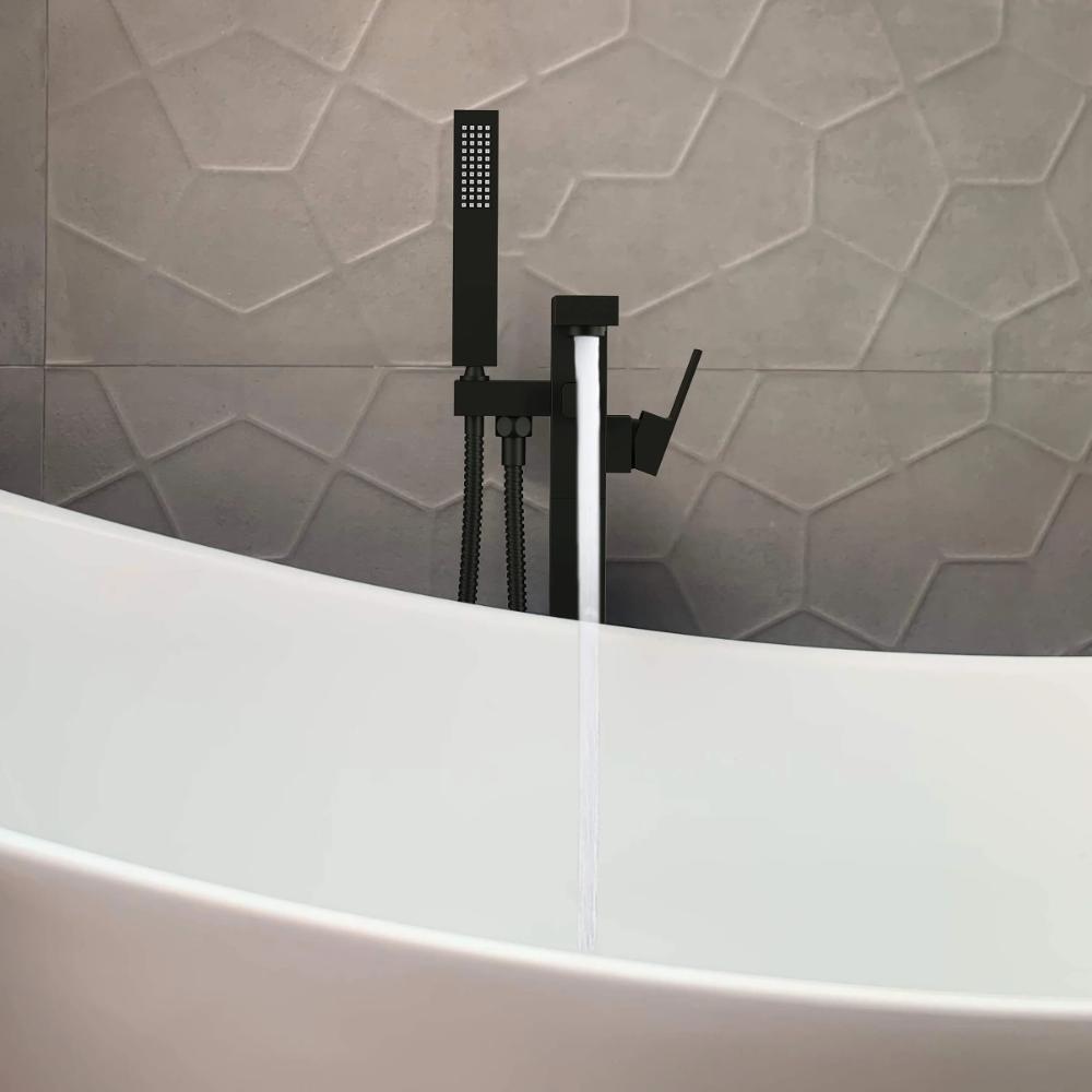 The Quadra Floor Mount Tub Filler With Hand Shower in black fills a white freestanding tub, set against a geometric textured gray tile wall.