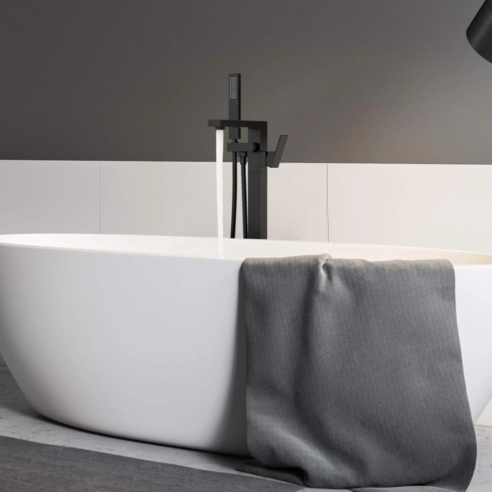 A modern white freestanding bathtub is paired with the Quadra Floor Mount Tub Filler With Hand Shower in sleek black. A gray towel rests on the tub’s edge, set against minimalist white and gray walls.