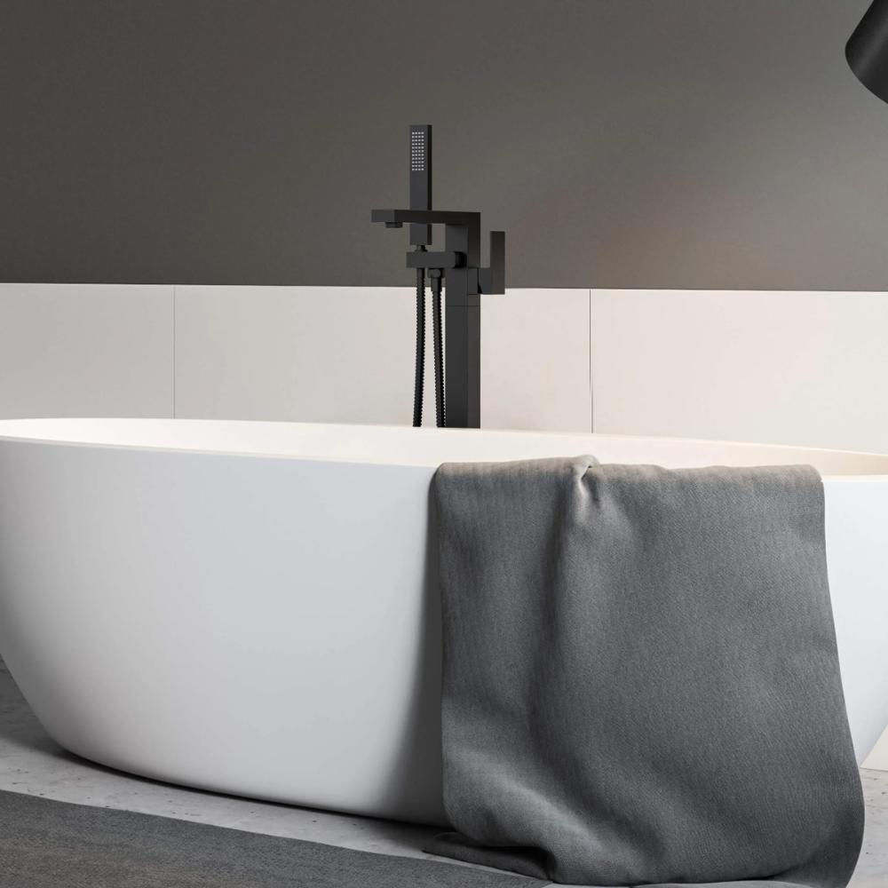 A modern white freestanding bathtub with a gray towel sits in a minimal bathroom featuring dark and light walls, complemented by the Quadra Floor Mount Tub Filler With Hand Shower in sleek black.