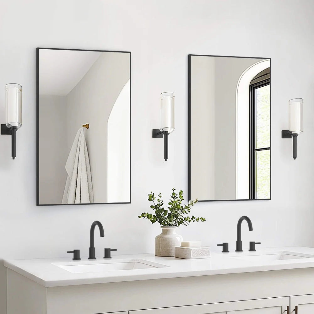 A modern bathroom showcases a double vanity with two Metal Framed Mirrors, black faucets, wall lights, a potted plant, and neatly stacked towels. In the background, a towel hangs on a gold hook.