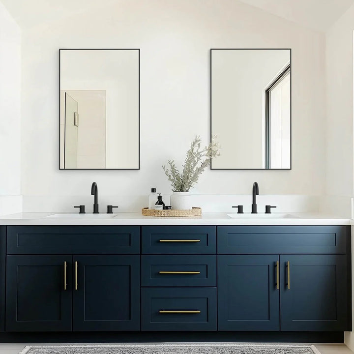 Modern bathroom with a dark blue double sink vanity, gold handles, two black faucets, and Metal Framed Mirrors above each sink. A decorative tray holding a plant and toiletries sits on the counter.
