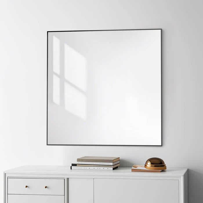 The Metal Framed Mirror with a slim black edge hangs on a white wall above a minimalist cabinet, reflecting sunlight and window shadows, while books and a bronze decor piece sit on the cabinet below.