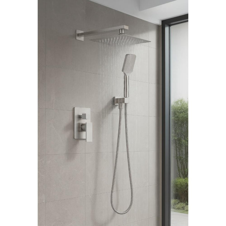 The Quadra Pressure Balanced Wall Mount Shower System features a large square overhead showerhead, a handheld attachment, and brushed metal controls, installed against light gray tiles beside a glass window.