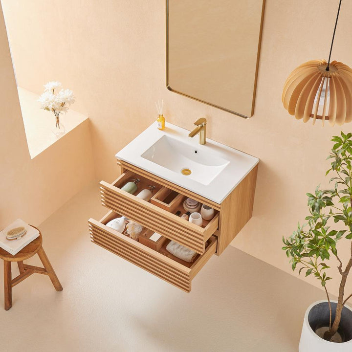 Minimalist bathroom featuring the Linea 30 Inch Wall Mounted Bathroom Vanity with a white sink, gold faucet, open drawers with toiletries, wall mirror, potted plant, wooden stool, pendant light, and a vase of white flowers on a ledge.