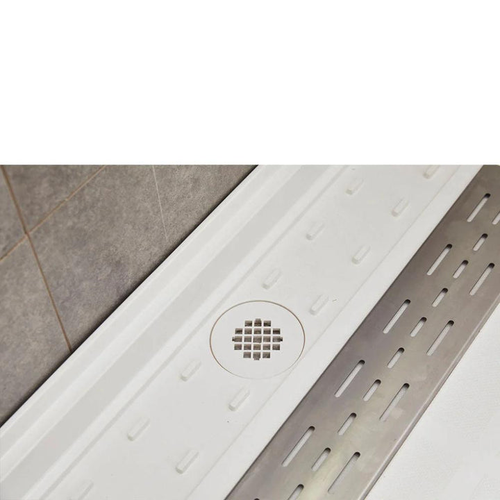 A close-up of the Bhavana Single Threshold Shower Base shows its white surface, central square drain, and a long rectangular silver grate on the right, with gray tiles on the adjacent wall.