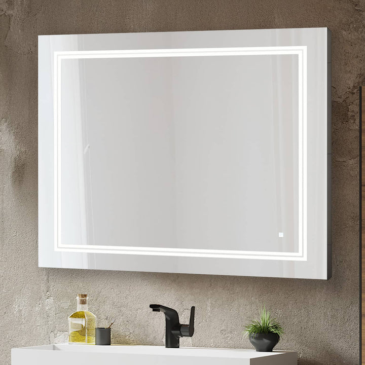 The Soho 36 Inch x 30 Inch LED Lighted Bathroom Mirror hangs above a white sink with a black faucet. On the counter are a bottle, glass, and small potted plant, all set against a textured wall.