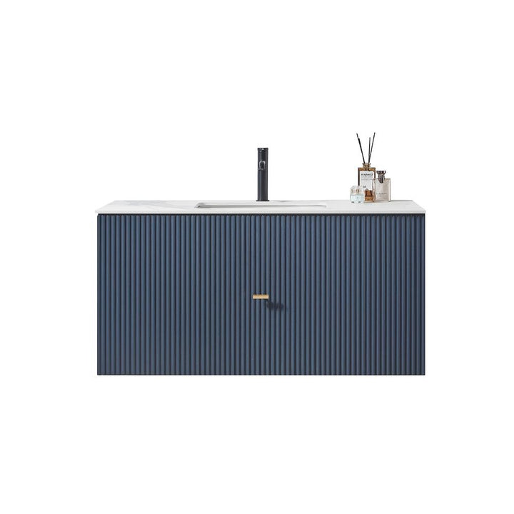 A modern Barcelona 42 Inch Wall Mount Bathroom Vanity features a ribbed navy blue cabinet, a white countertop, a black faucet, and decorative items like a reed diffuser and glass containers.