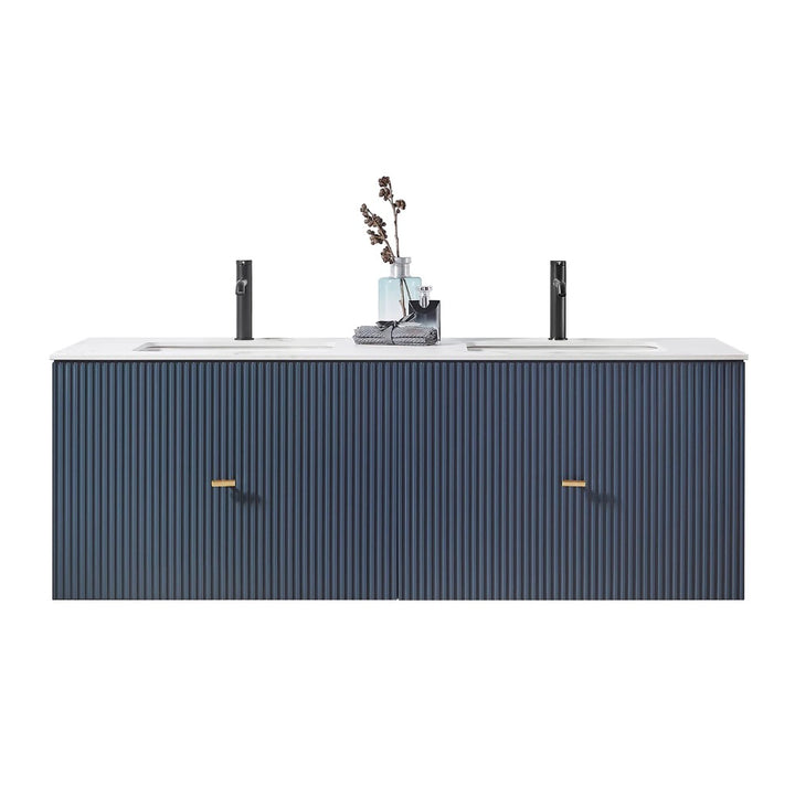 The Barcelona 60 Inch Wall Mount Double Bathroom Vanity With Top features a navy blue, vertically grooved front, gold handles, and two black faucets. The countertop is accented with decorative vases and a folded towel.