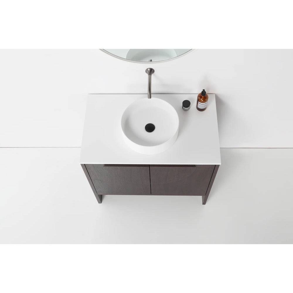 Overhead view of the Celle 36 Inch Bathroom Vanity With Vessel Sink, showcasing a round white sink, minimalist faucet, two brown cabinet doors, a small bottle, and black cup on its white countertop beneath a partial circular mirror.