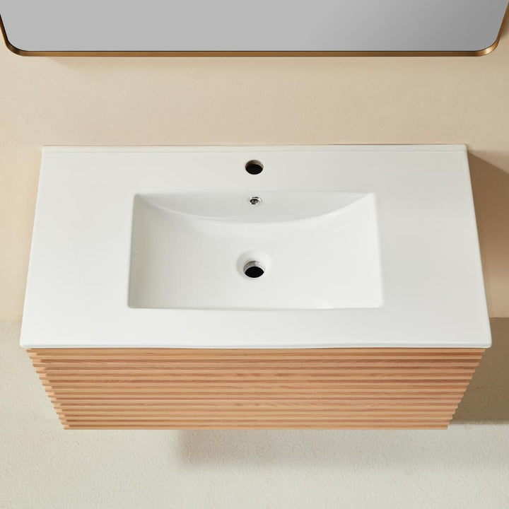 A rectangular white sink with a single faucet hole rests in the Linea 36 Inch Wall Mounted Bathroom Vanity with light wood slats, shown from above against beige walls and flooring.