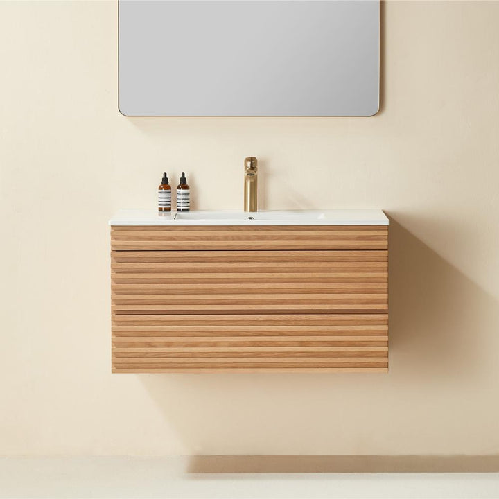 A minimalist Linea 36 Inch Wall Mounted Bathroom Vanity with a wood-grain finish, white countertop, modern gold faucet, two small bottles, and a large rectangular mirror above, set against a beige wall.
