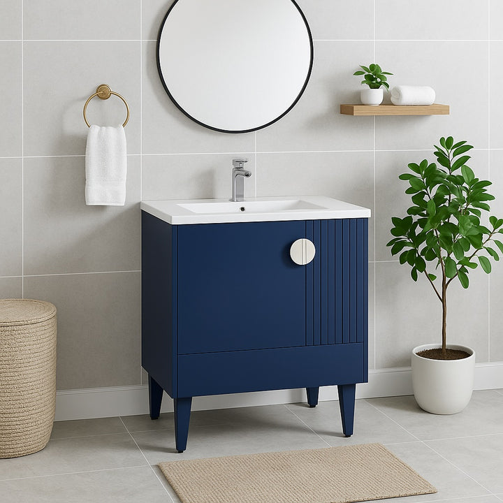 A modern bathroom featuring a Venezian 30 inch Bathroom Vanity With Sink, white countertop, round mirror, wall towel shelf, gold hand towel ring, potted plant, woven basket, and beige floor mat on light gray tiled walls and floor.