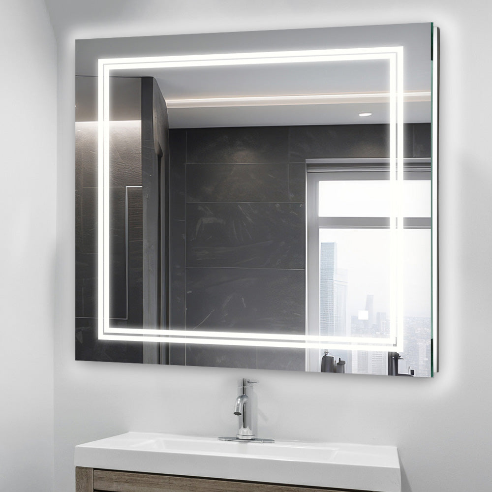 A modern bathroom features the Soho 30 Inch x 30 Inch LED Lighted Bathroom Mirror above a sink and wooden vanity, its edge-lit LEDs glowing as a window reflects a cityscape in the background.