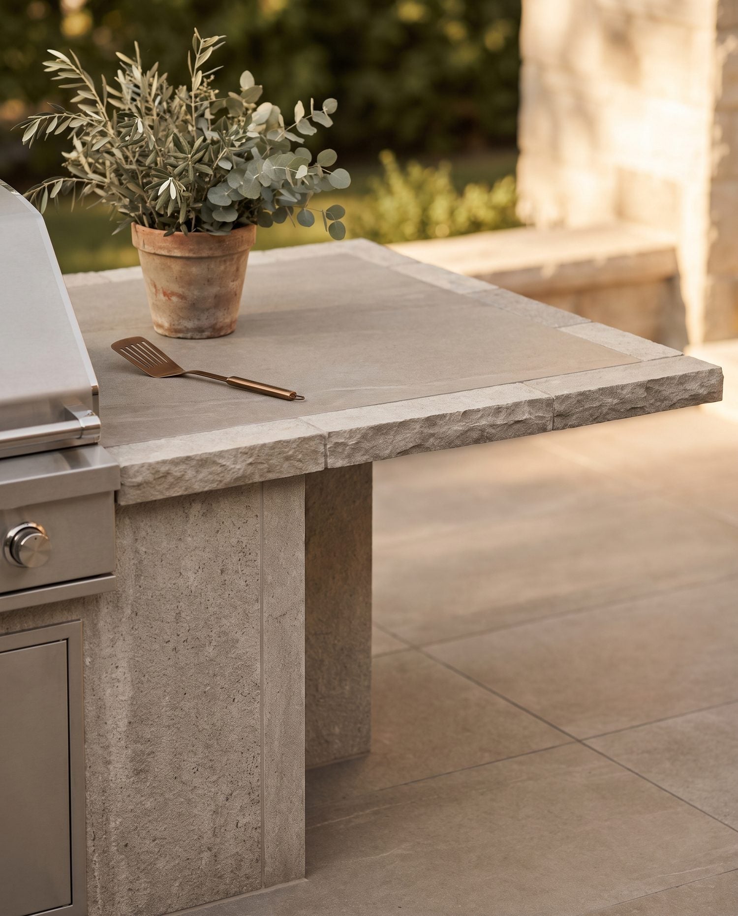 Styled corner of an outdoor kitchen showing a grill island beside a porcelain paver patio and slab counter, golden-hour light