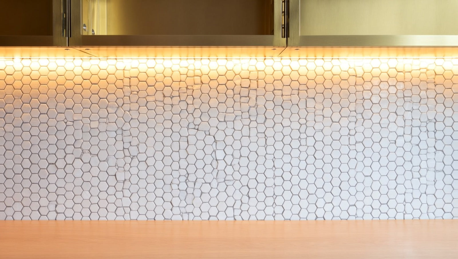 Matte white hexagon mosaic backsplash with charcoal grout beneath brass shelving and warm under-cabinet lighting.