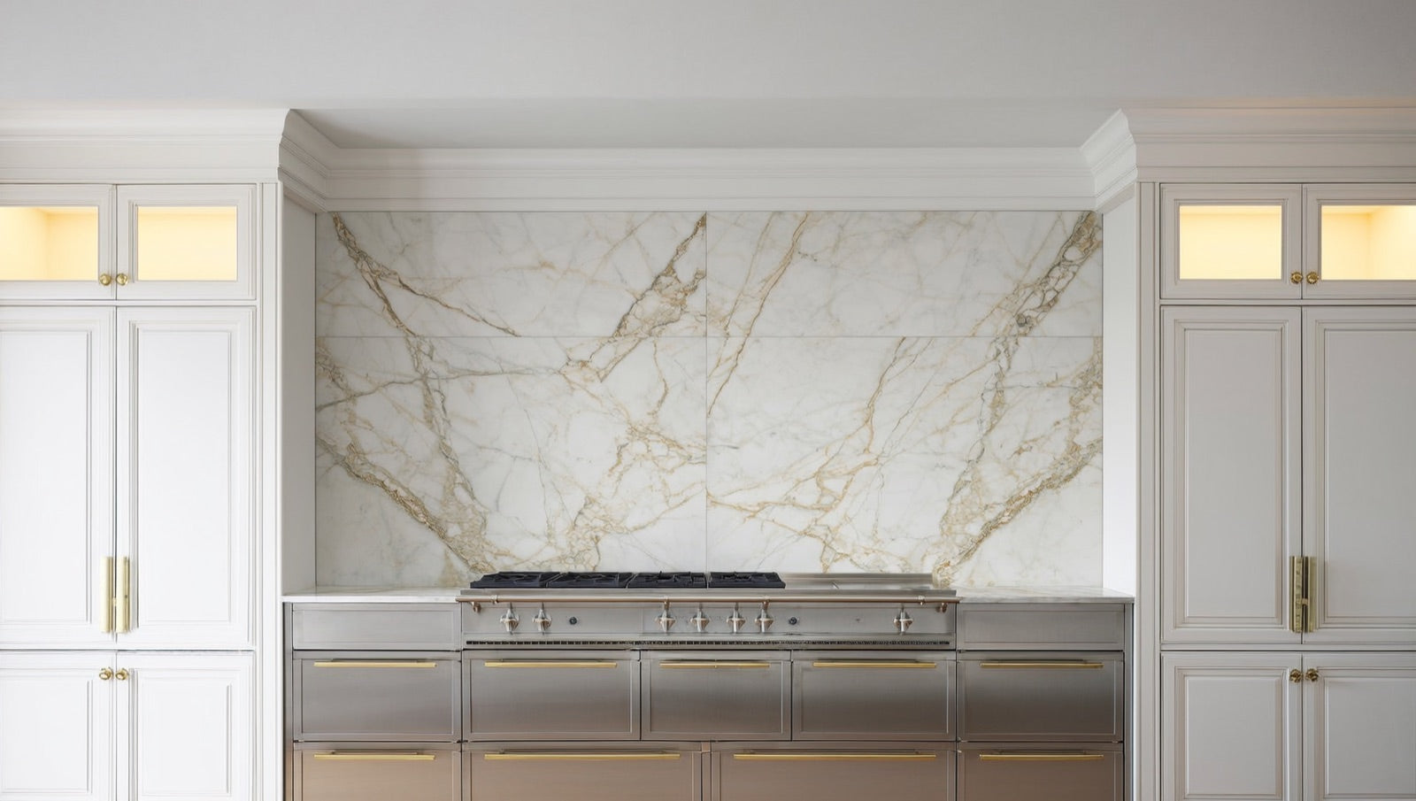 Marble-look porcelain tile with Calacatta-style grey and gold veining covering a kitchen range wall between white cabinets.
