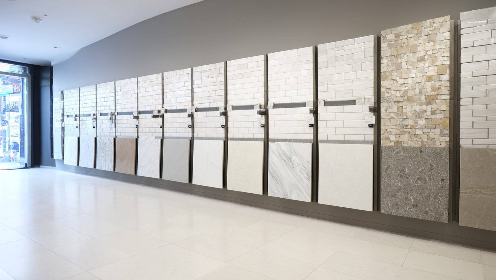 Tile showroom display wall with upright panels of subway tile, marble-look porcelain, and mosaic sample boards in whites, beiges, and greys.
