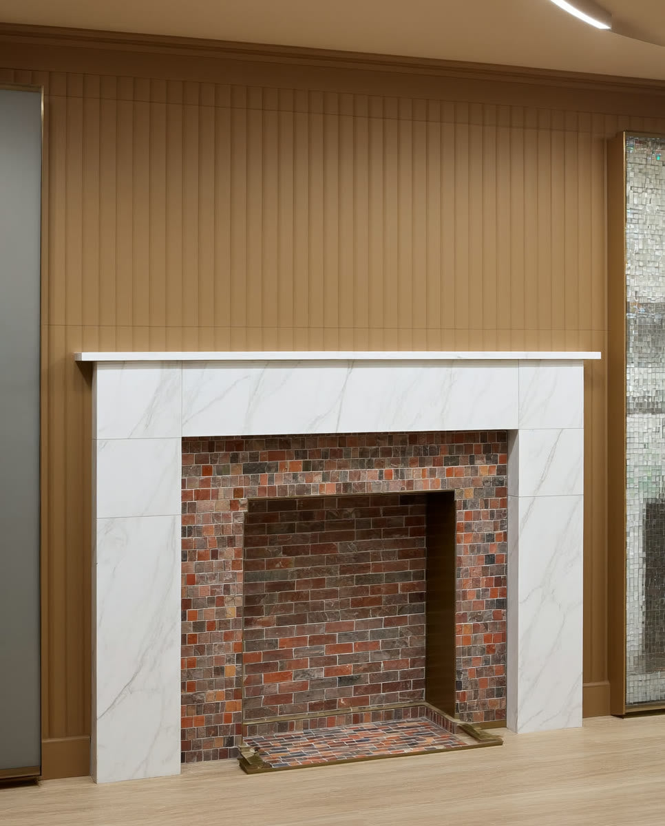 A corner fireplace clad in large-format marble-look tile with a stone hearth, styled with a vase and books against cream plaster walls in the ANVE style.