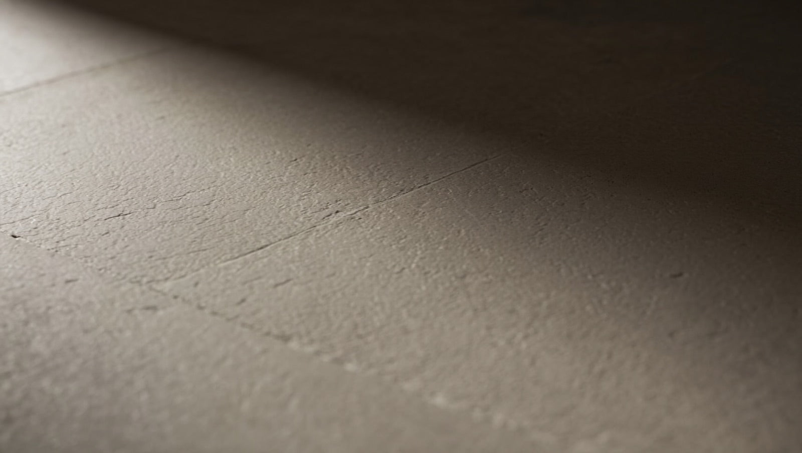 Close-up of a matte stone-look porcelain floor tile surface in warm grey, with low raking light revealing its fine, even micro-texture