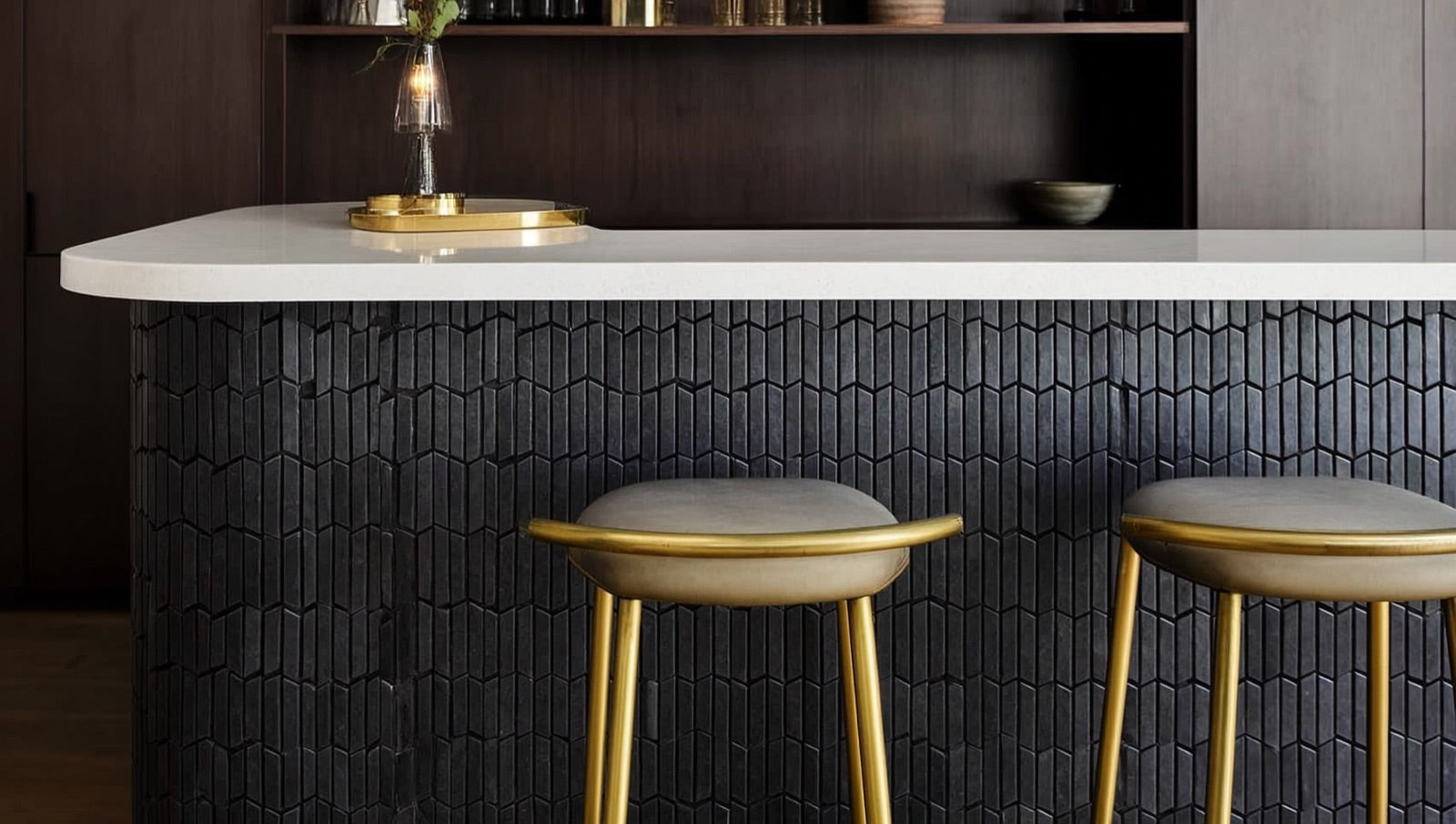 Home bar front clad in vertically stacked matte black hexagon mosaic tile beneath a white countertop, with brass-footed bar stools and pendant light grazing the honeycomb surface.
