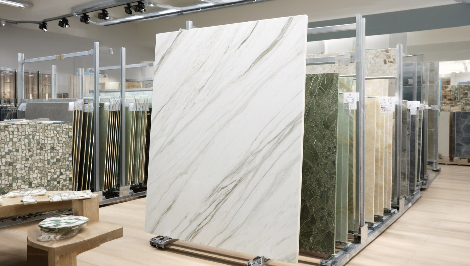 Showroom display of full-size porcelain slabs standing upright in steel viewing racks, a polished white marble-look panel in front of onyx-green and travertine-beige slabs, with mosaic sample boards nearby.