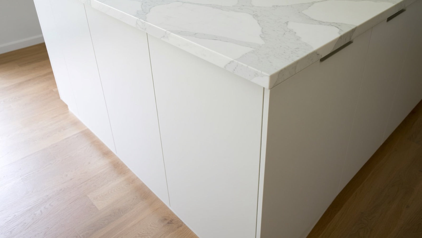 Corner of a kitchen island clad in white marble-look porcelain slab, grey veining flowing continuously from the top over the mitered edge and down the vertical end panel to the oak floor.