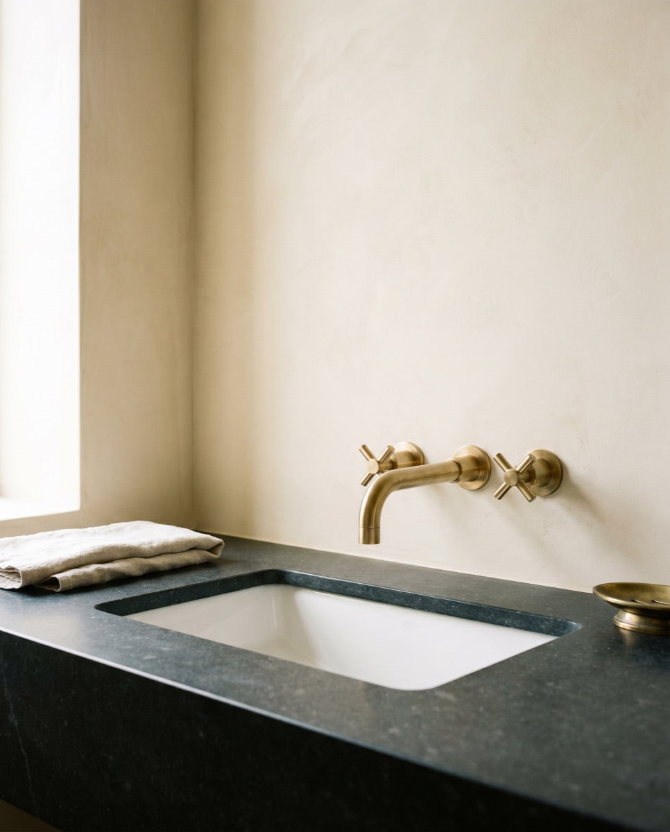 An undermount white ceramic bathroom sink in a honed stone vanity with a brushed-brass faucet in the ANVE showroom.