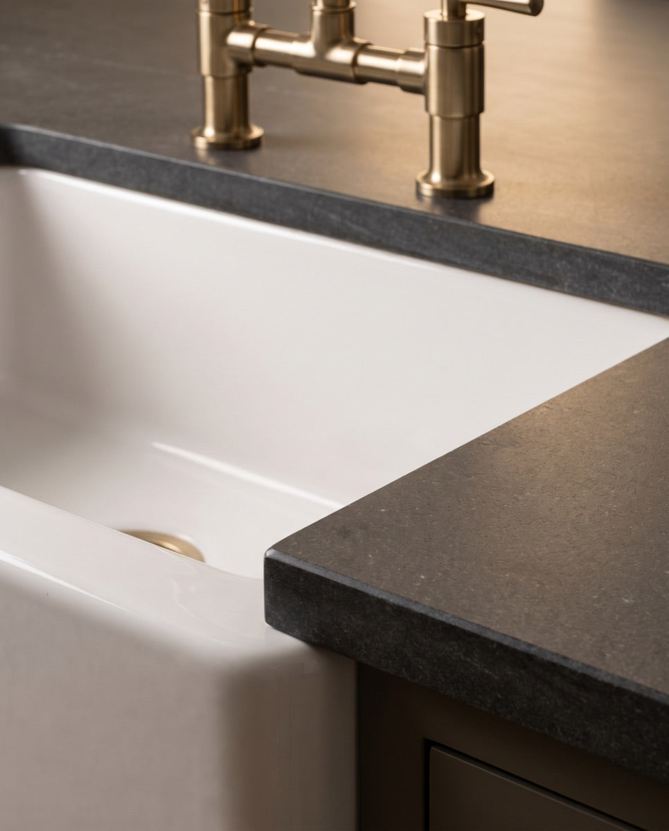 A detail of a white fireclay farmhouse sink apron meeting a honed stone counter with a brushed-brass faucet.