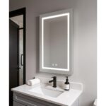 A modern bathroom vanity features the Daytona 24 Inch x 30 Inch LED Lighted Bathroom Mirror above a white sink with black faucet, rolled towel, and soap dispenser on a light gray countertop, with a black door and tiled shower in the background.