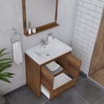 Wood vanity with a white sink and open cabinet doors, revealing folded towels inside. A wall mirror, toothbrush holder, soap, and towel ring are visible. The bathroom has gray tile flooring and white subway tile walls.