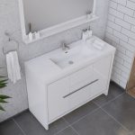 Modern bathroom vanity with a white sink, silver faucet, and storage cabinets below. Towels, toiletries, and a mirror are arranged neatly; the walls are tiled and the floor has large gray tiles.