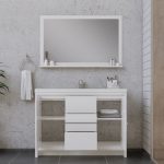 Modern bathroom with a large rectangular mirror above a white vanity with open shelves and drawers; towels and toiletries are neatly arranged, and a towel hangs on a ring to the left. The wall is light gray concrete.