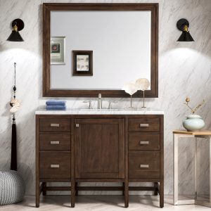 A modern bathroom showcases an Addison 48 vanity in Mid Century Acacia with a marble countertop and large mirror. Dark sconces flank the mirror. Decor features framed leaf art, a blue towel, and elegant accessories amid white textured walls and tiled accents.