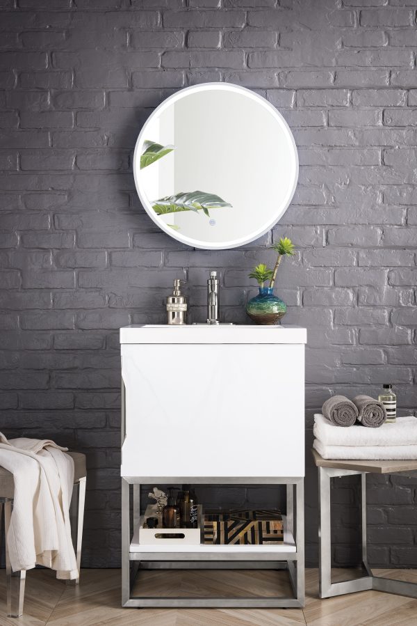 A modern bathroom displays the Columbia 24 Bathroom Vanity in Glossy White with Brushed Nickel fixtures and a round mirror above it, set against a gray brick wall. A plant and toiletries decorate the sink, while a nearby stool holds folded towels on a light wood-patterned floor.