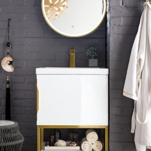 A stylish bathroom interior with a round mirror, Columbia 24 Bathroom Vanity in Glossy White and Radiant Gold, and a dark gray textured wall. Open shelves hold rolled towels and decor, while a woven stool and modern light fixture enhance elegance. A white robe hangs nearby.