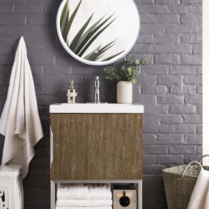 In a bathroom, the Columbia 24 Bathroom Vanity Cabinet in Latte Oak with Brushed Nickel supports a sleek white sink. A round mirror decorated with leaf designs hangs above it, while a towel and woven basket sit nearby against the gray-painted brick wall.
