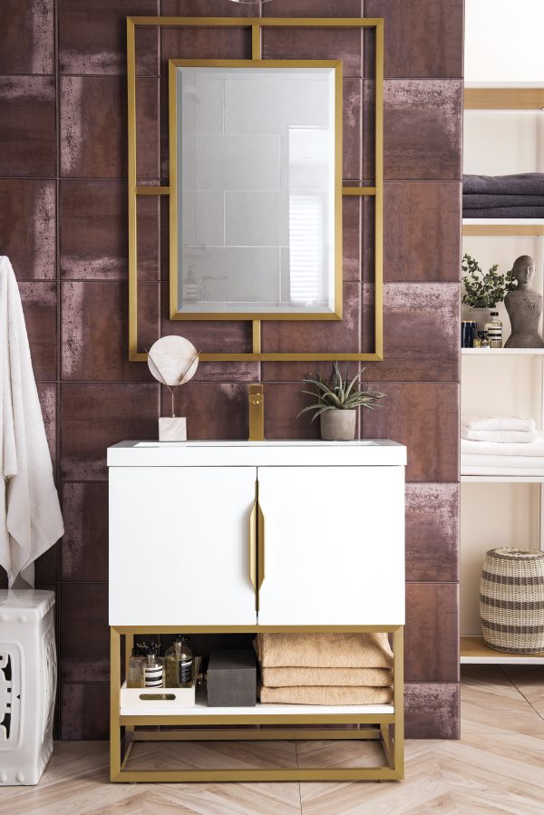 The elegant bathroom features the Columbia 31.5 Bathroom Vanity in Glossy White with Radiant Gold accents. A gold-framed rectangular mirror hangs above, while shelves with folded towels and decor complement the warm, sophisticated ambiance of richly tiled brown walls.
