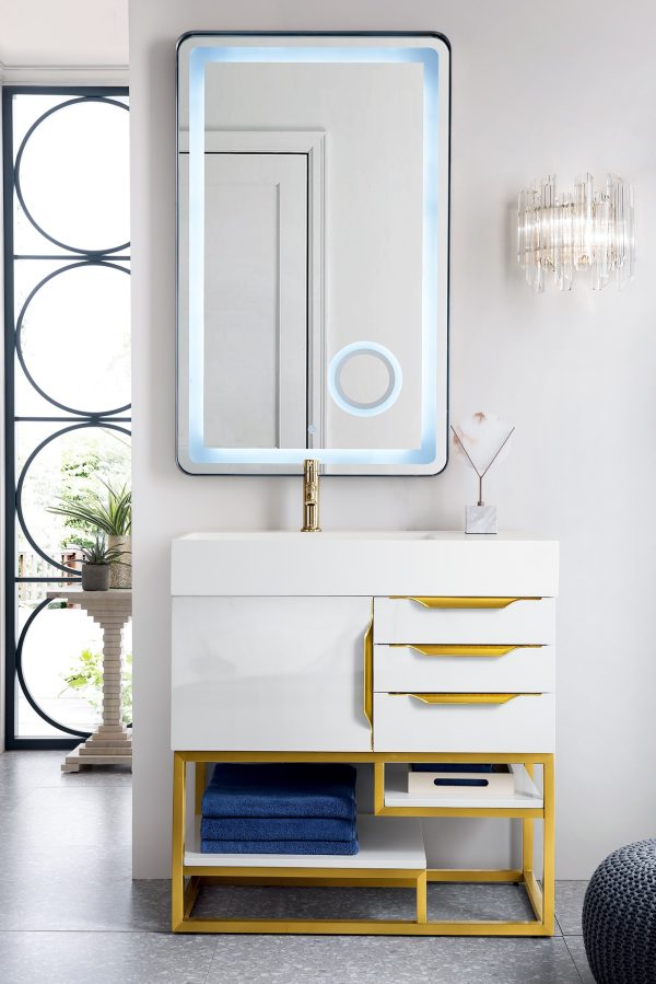 A modern bathroom showcases the Columbia 36 Single Bathroom Vanity in Glossy White and Radiant Gold, with a backlit rectangular mirror above. It includes three drawers, a shelf with blue towels, and a pristine tray. A stylish wall sconce is on the right; circular window patterns adorn the left.