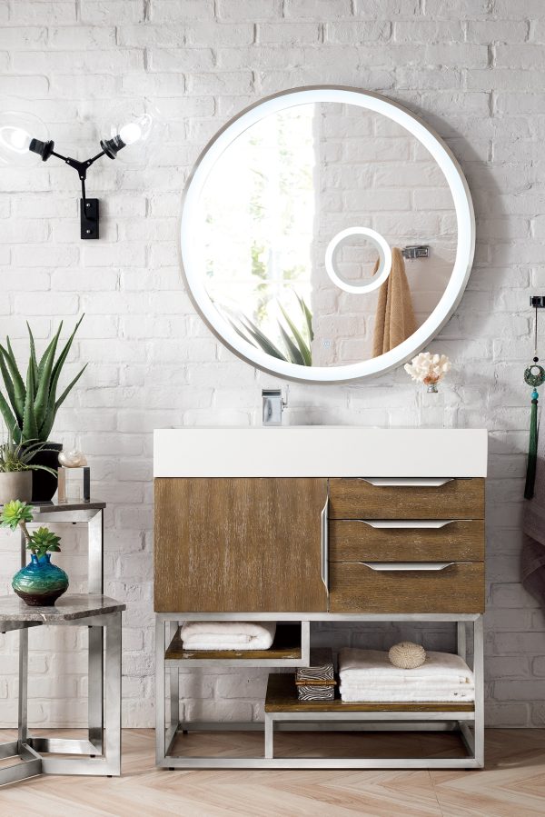 A modern bathroom showcases a Columbia 36 Single Bathroom Vanity in Latte Oak with Brushed Nickel hardware. The vanity features drawers and open shelves for towels and decor. White brick walls, a small table with plants, an illuminated round mirror, and wall-mounted light complete the contemporary design.