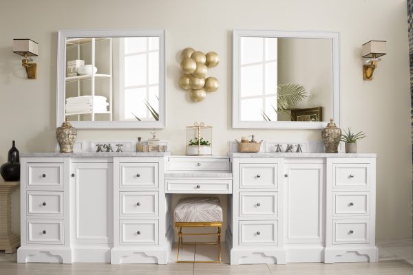 The De Soto 118 Double Bathroom Vanity in Bright White with Arctic Solid Surface Top includes two mirrors and gold faucets. Decorated with vases, a small plant, and a gold wall sculpture, it also features a cleverly placed stool under the center countertop for convenience.