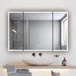 A modern bathroom features a round stone sink on a wooden counter, folded towels, and toiletries. An Edge Royal 48 Inch LED Lighted Mirror 3 Door Medicine Cabinet serves as a large mirror, reflecting the shower with gray tiles and a window.