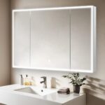 A modern bathroom features the Edge Royal 48 Inch LED Lighted Mirror 3 Door Medicine Cabinet above a white sink, with a chrome faucet, a small plant, and towels on the countertop against a beige wall.